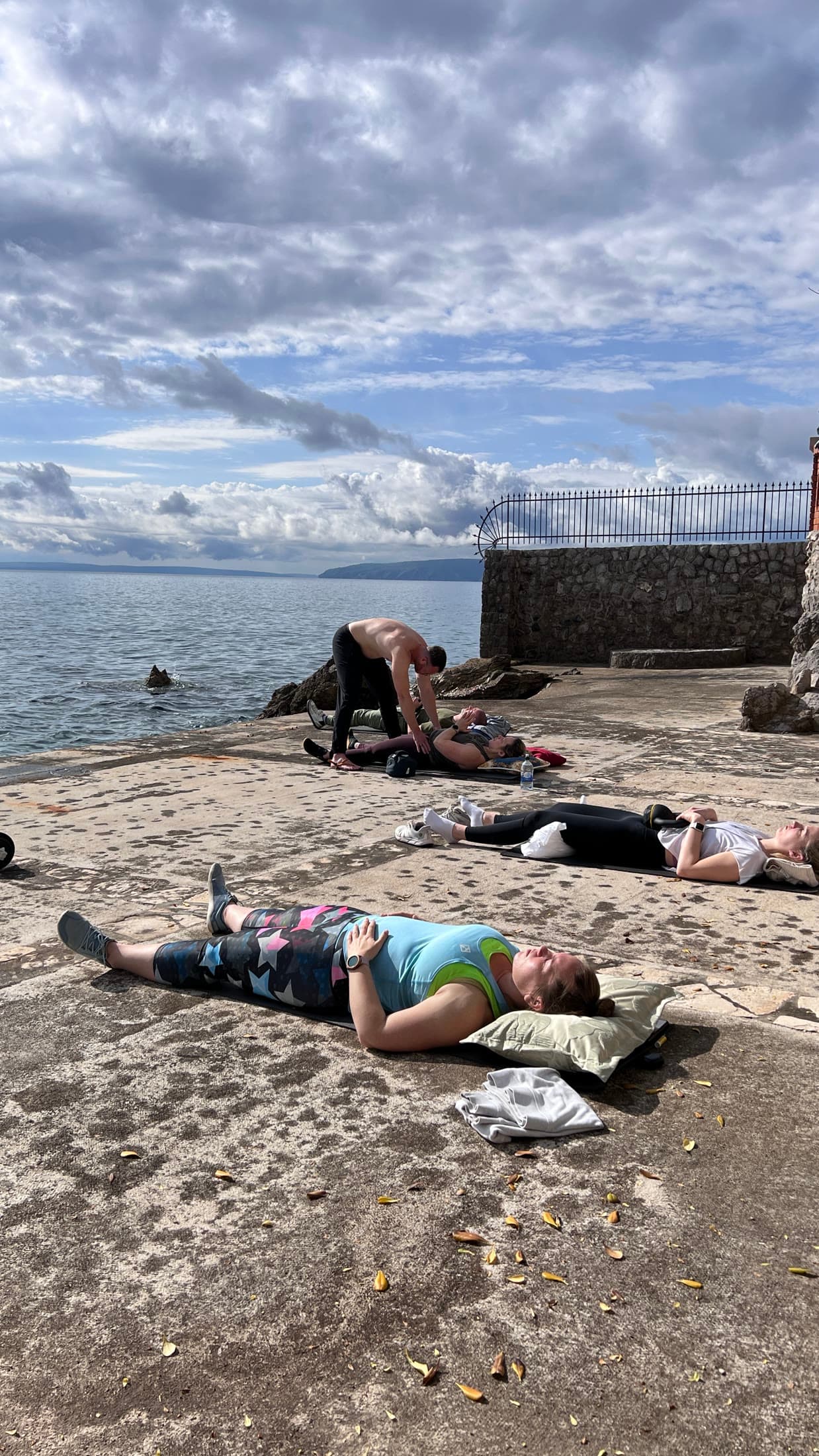 A participant lying on a stone terrace by the sea during breathwork, with hands-on guidance in the background