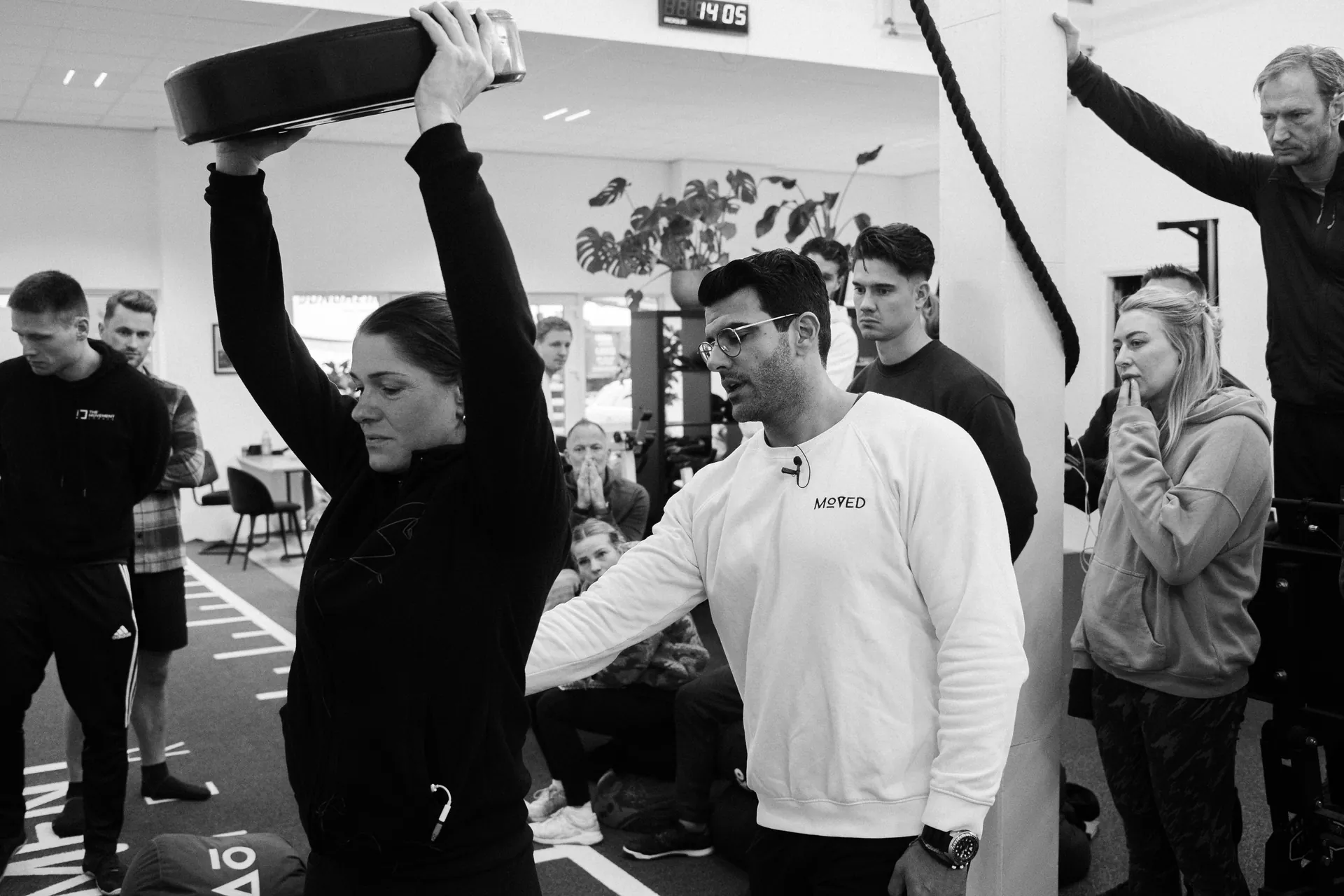A coach pressing a weight plate overhead while Richard Aceves spots the lockout